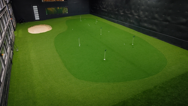 Plaša iekštelpu golfa ripināšanas zona ar vairākiem karodziņiem un smilšu bunkuru